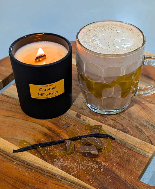 A wooden board with chocolate shavings, a vanilla bean and a milkshake with a lit wooden wick candle in a matte black jar