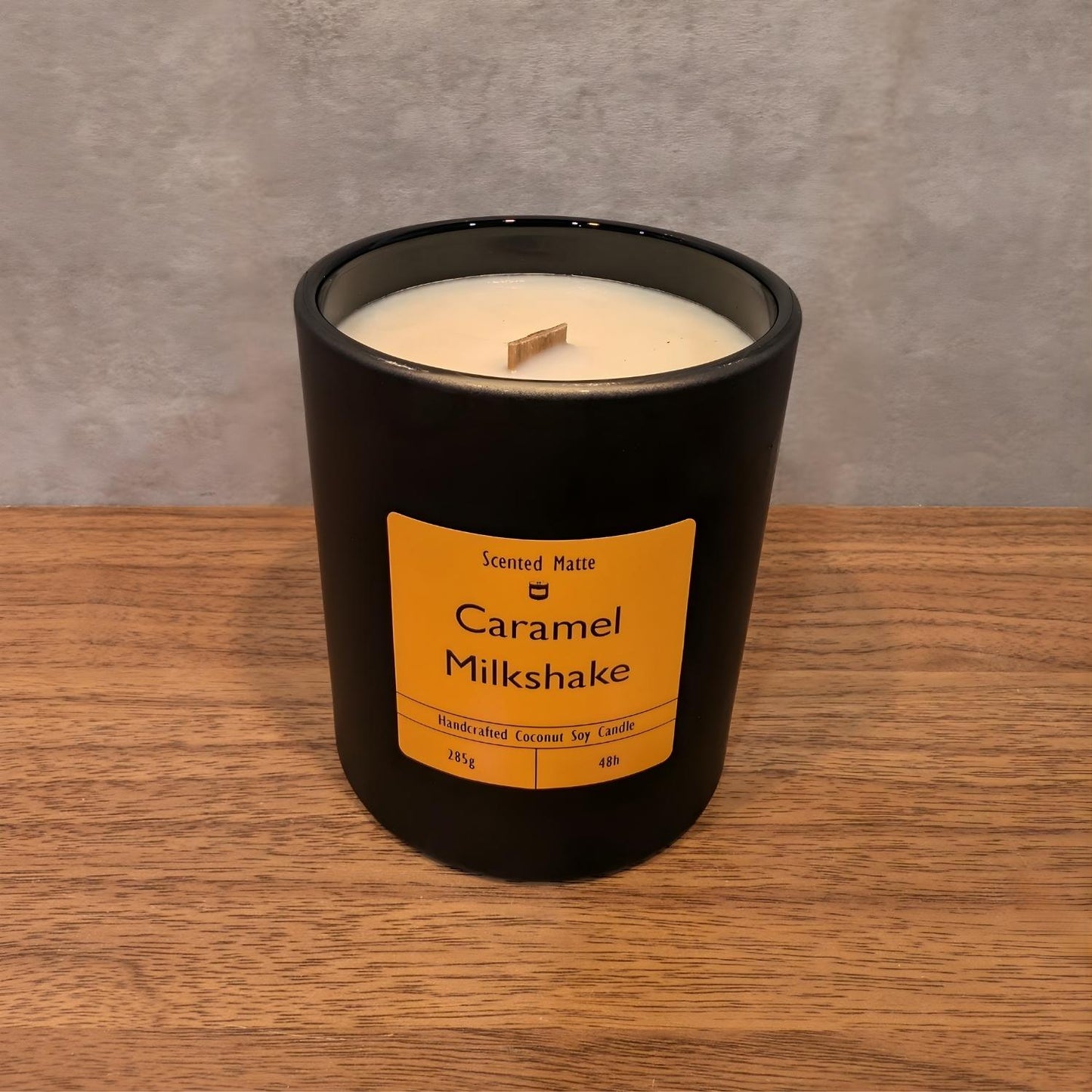 A minimalist matte black candle jar with a wooden wick, sitting on a light oak wood surface against a textured grey background.