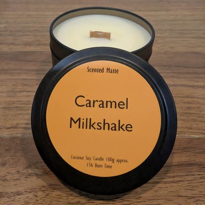 A wooden wick candle in a black tin with orange label on the lid on a wooden surface.