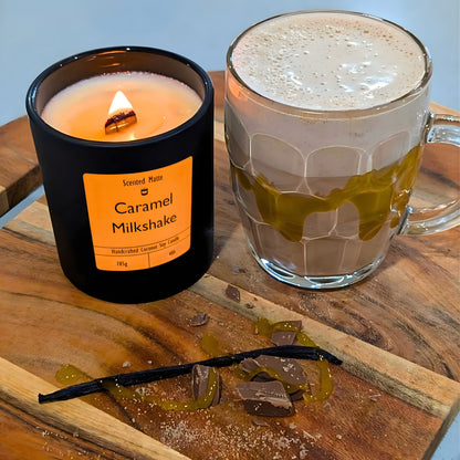 A minimalist matte black candle jar with a lit wooden wick, placed next to a foamy milkshake with golden caramel sauce and a whole vanilla bean pod on a wooden board.