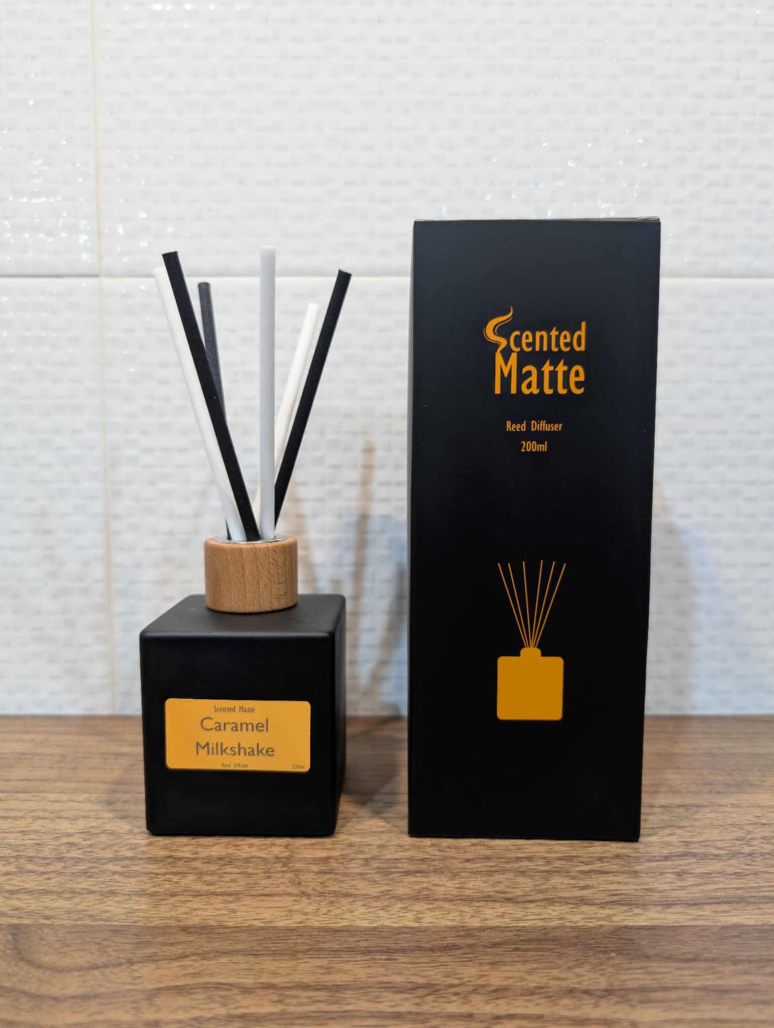 Matte black diffuser bottle with black and white reed s and wooden cap, next to its packaging on a wooden surface.icks