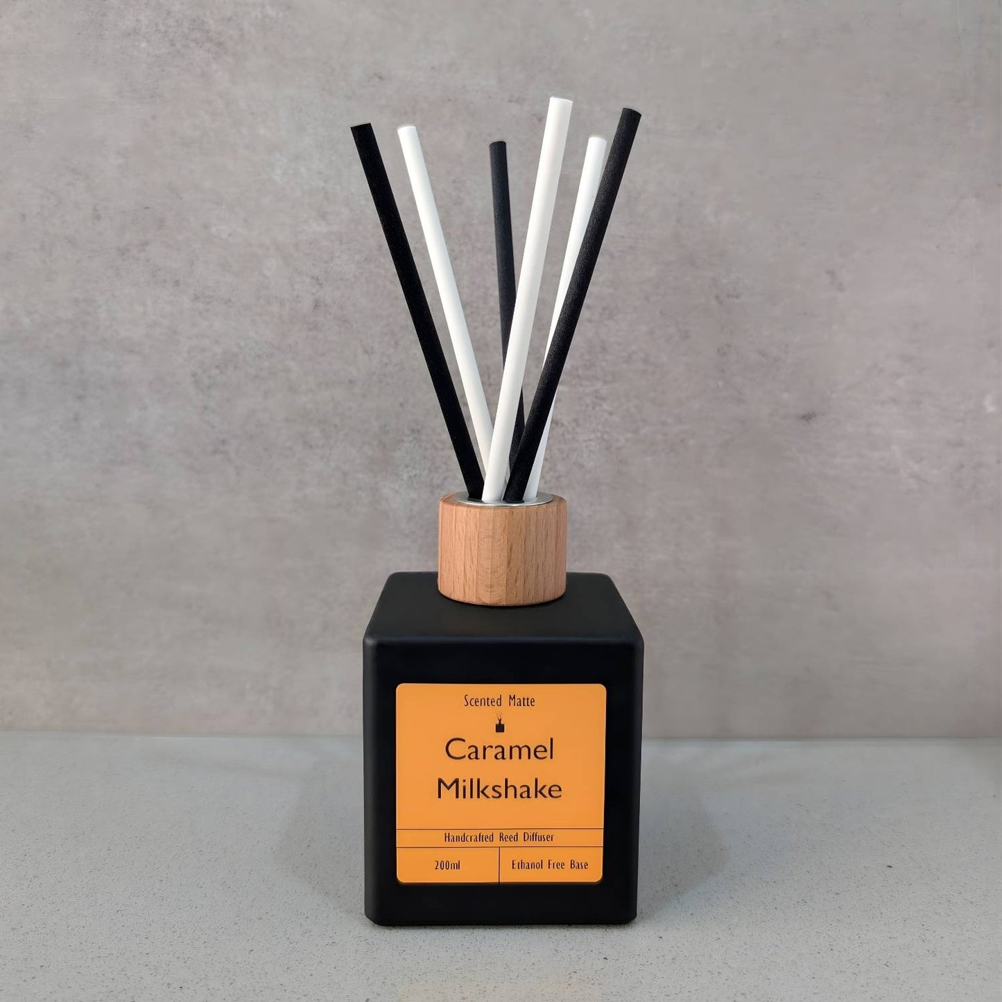 A minimalist matte black square reed diffuser with an orange label, light wood cap and six black/white fiber reeds, set against a grey background.