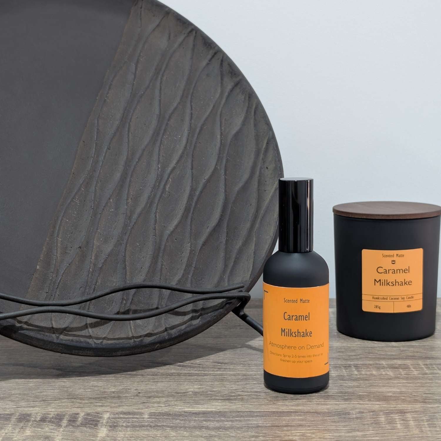 A matte black room spray bottle and a minimalist matte black candle jar, styled next to a large textured charcoal grey decorative plate.