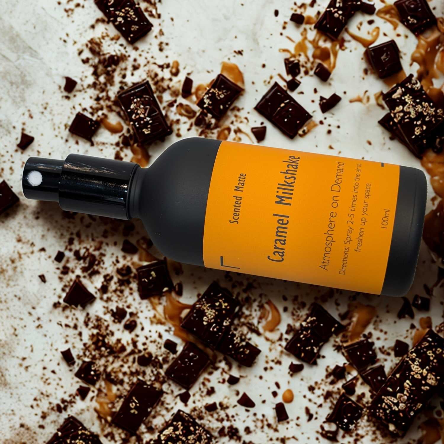 A minimalist matte black room spray bottle with a vibrant orange label, placed amoung golden caramel sauce and chocolate chunks.