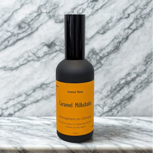 A minimalist matte black 100ml room spray bottle with a vibrant orange label, centered on a white marble surface.