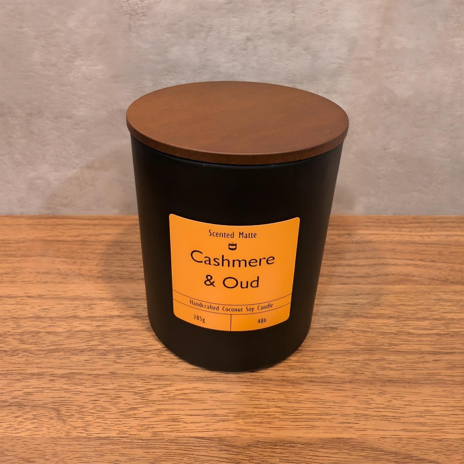 A minimalist matte black candle jar with a vibrant orange label and a dark wood lid.