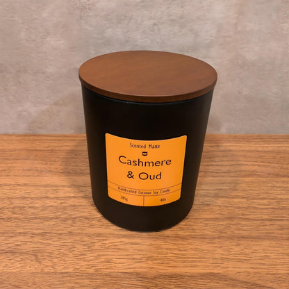 A minimalist matte black candle jar with a vibrant orange label and a dark wood lid.