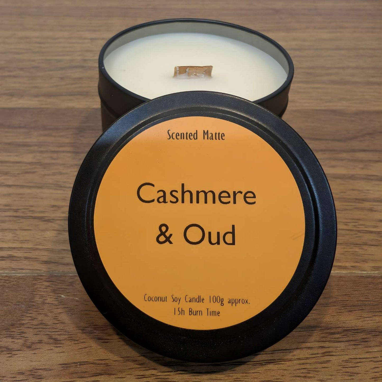 A wooden wick candle in a black tin with orange label on the lid on a wooden surface.