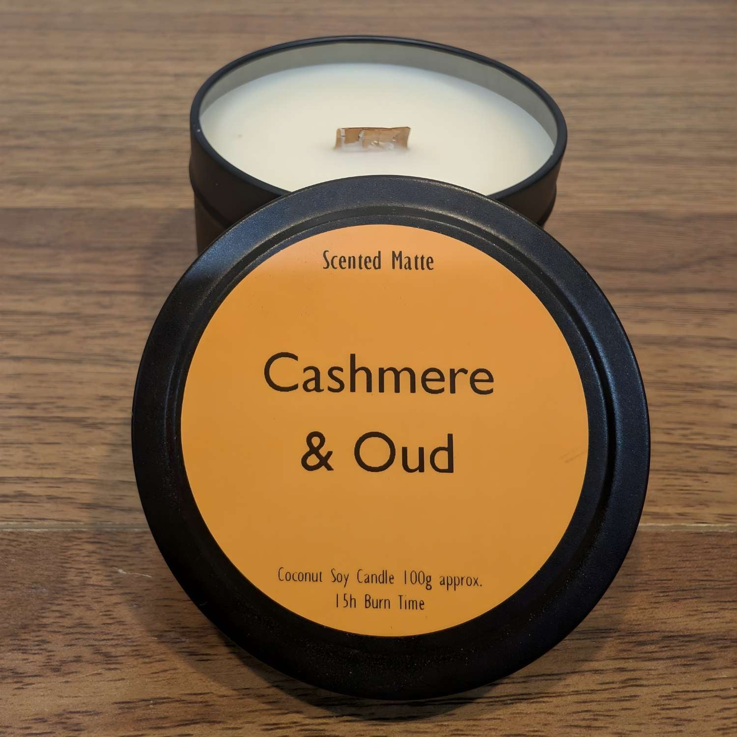 A wooden wick candle in a black tin with orange label on the lid on a wooden surface.
