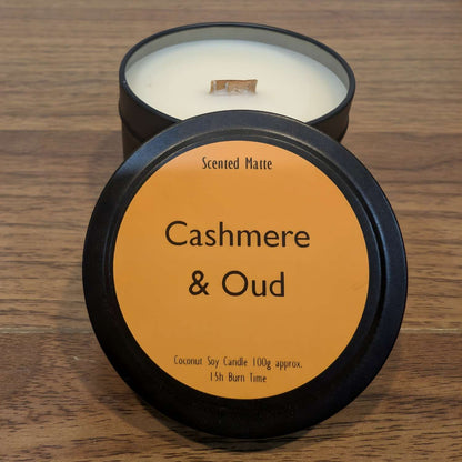 A wooden wick candle in a black tin with orange label on the lid on a wooden surface.
