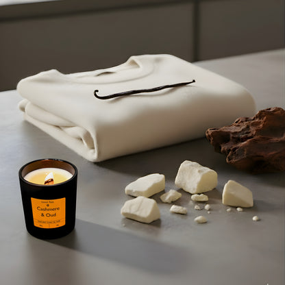 A minimalist matte black candle jar with a lit wooden wick on a concrete bench, next to a soft cream knitted fabric with dark oud wood and cocoa butter chunks to the side.