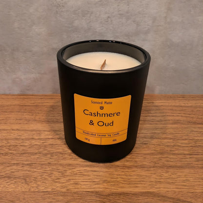 A minimalist matte black candle jar with a wooden wick, sitting on a light oak wood surface against a textured grey background.