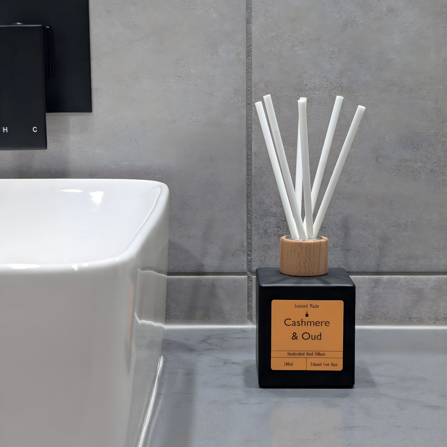 A minimalist matte black reed diffuser with orange label, wooden cap and white fiber reeds, sitting on a grey stone vanity next to a white ceramic sink and grey tiled wall.