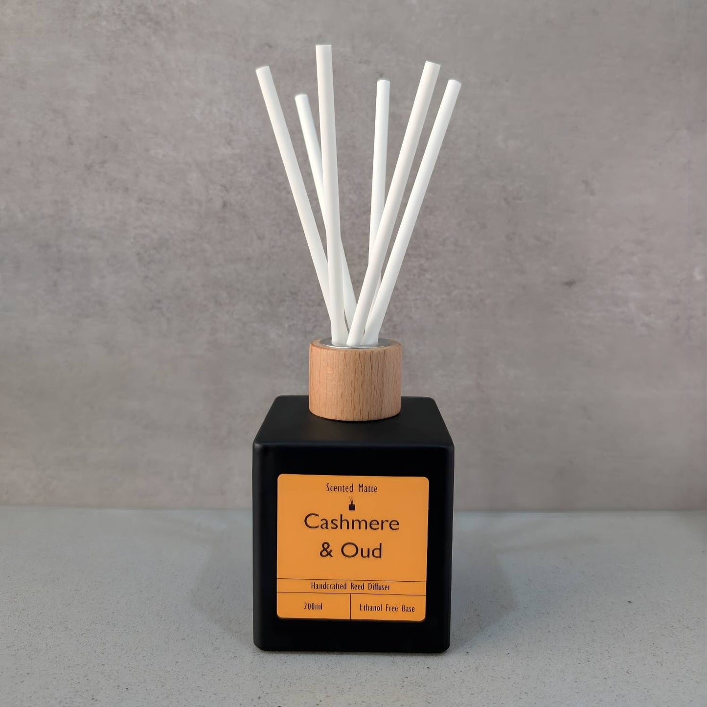 A minimalist matte black square reed diffuser with an orange label, light wood cap and six white fiber reeds, set against a grey background.