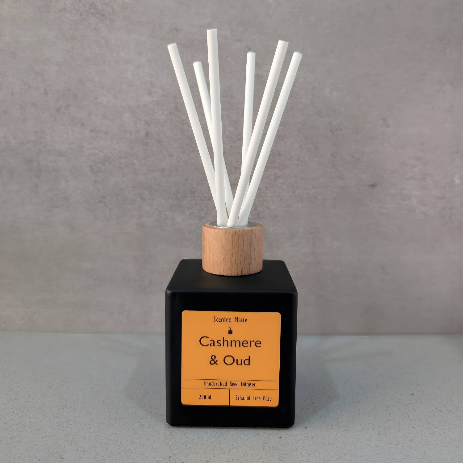 A minimalist matte black square reed diffuser with an orange label, light wood cap and six white fiber reeds, set against a grey background.