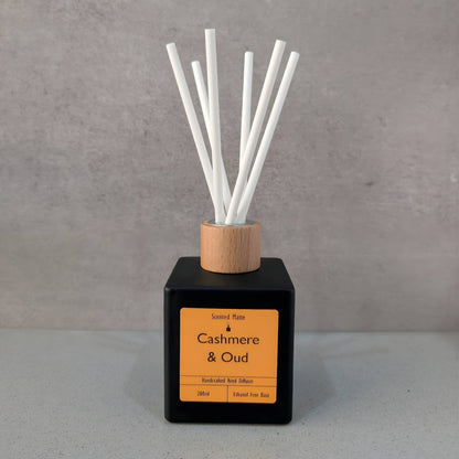 A minimalist matte black square reed diffuser with an orange label, light wood cap and six white fiber reeds, set against a grey background.