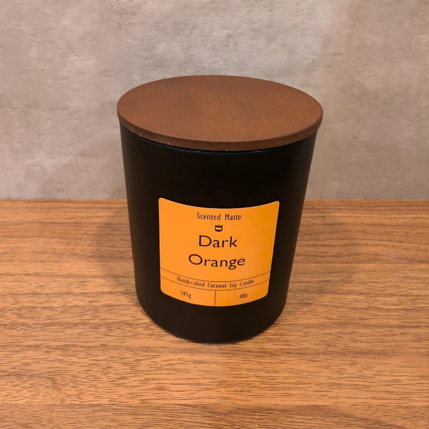 A minimalist matte black candle jar with a vibrant orange label and a dark wood lid.