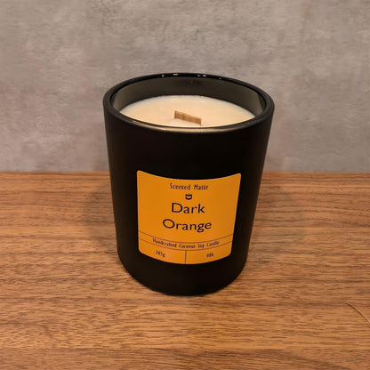 A minimalist matte black candle jar with a wooden wick, sitting on a light oak wood surface against a textured grey background.