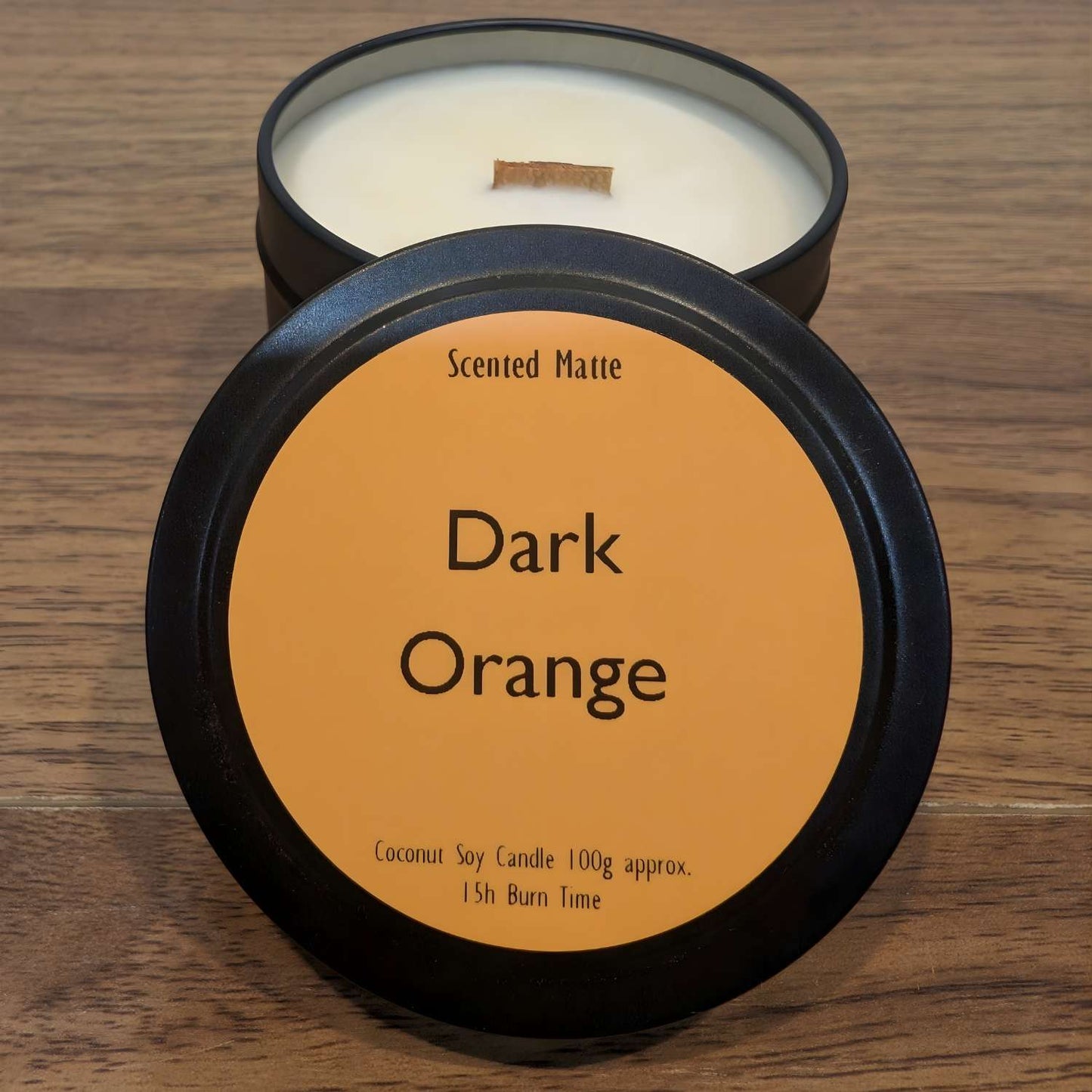 A wooden wick candle in a black tin with orange label on the lid on a wooden surface.