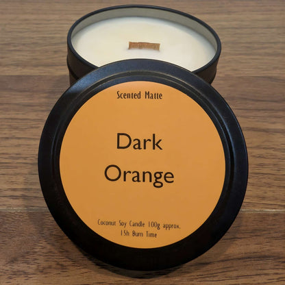 A wooden wick candle in a black tin with orange label on the lid on a wooden surface.