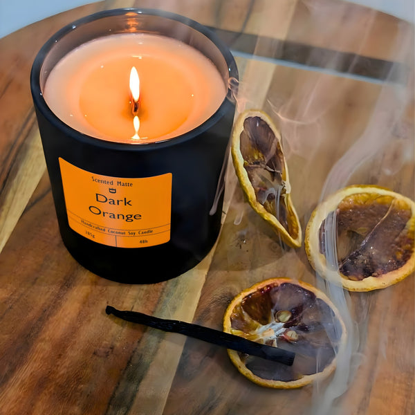 A minimalist matte black candle jar with a lit wooden wick, styled with dehydrated orange slices and a vanilla pod with whisps of smoke on a wooden board.
