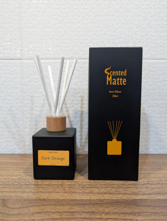 Matte black diffuser bottle with white reed sticks and wooden cap, next to its packaging on a wooden surface.