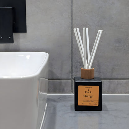 A minimalist matte black reed diffuser with orange label, wooden cap and white fiber reeds, sitting on a grey stone vanity next to a white ceramic sink and grey tiled wall.
