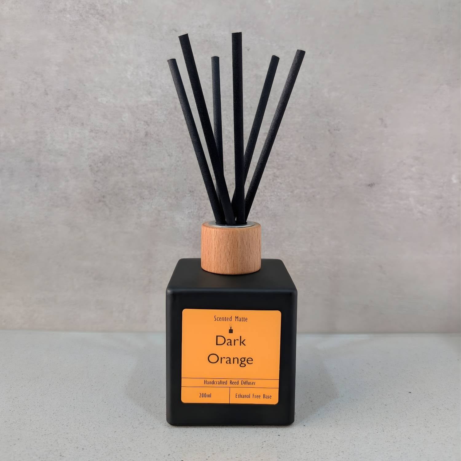 A minimalist matte black square reed diffuser with an orange label, light wood cap and six black fiber reeds, set against a grey background.