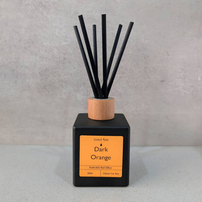 A minimalist matte black square reed diffuser with an orange label, light wood cap and six black fiber reeds, set against a grey background.