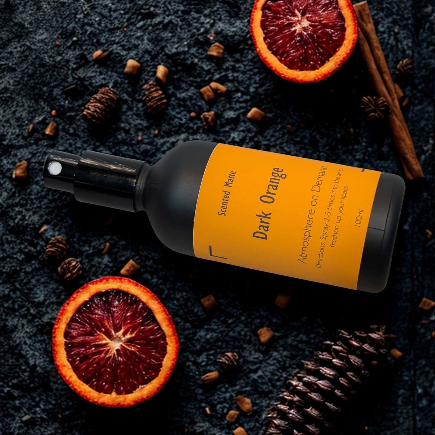 A minimalist matte black room spray bottle with a vibrant orange label, surrounded by dehydrated orange slices and whole pinecone.