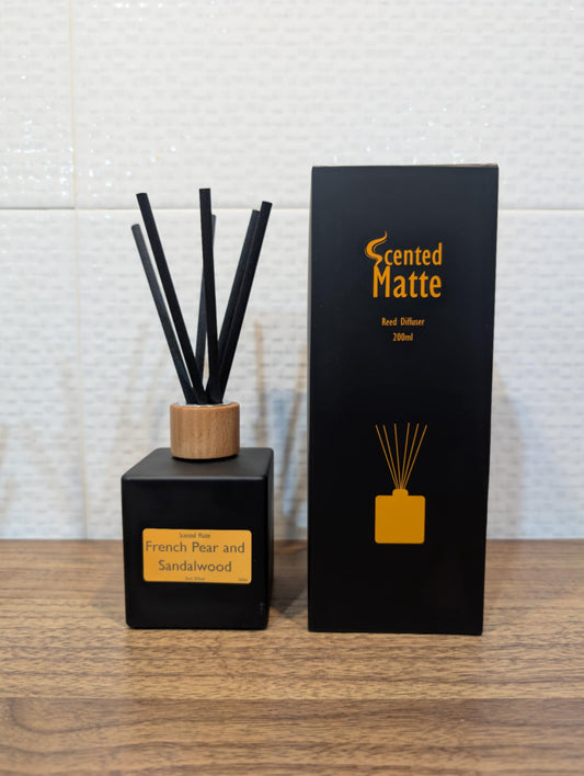 Matte black diffuser bottle with black reed sticks and wooden cap, next to its packaging on a wooden surface.