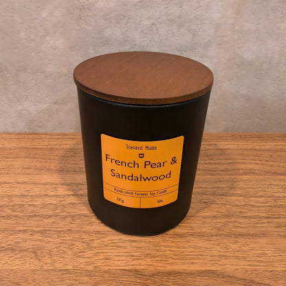 A minimalist matte black candle jar with a vibrant orange label and a dark wood lid.