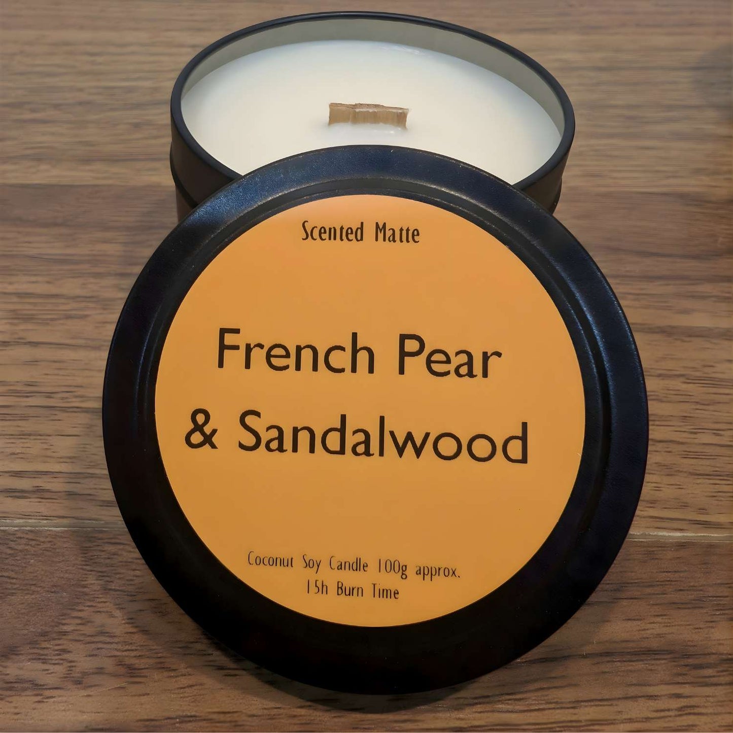 A wooden wick candle in a black tin with orange label on the lid on a wooden surface.