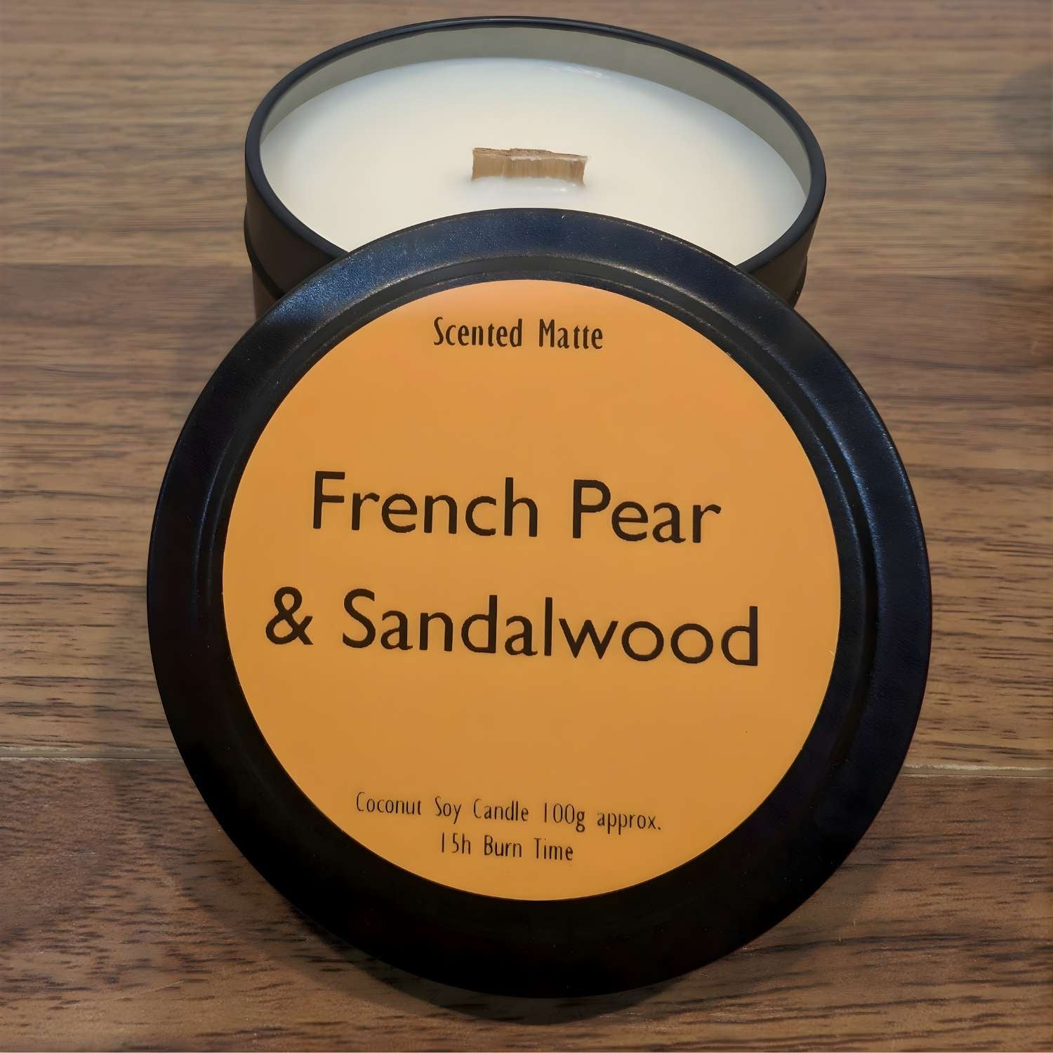 A wooden wick candle in a black tin with orange label on the lid on a wooden surface.
