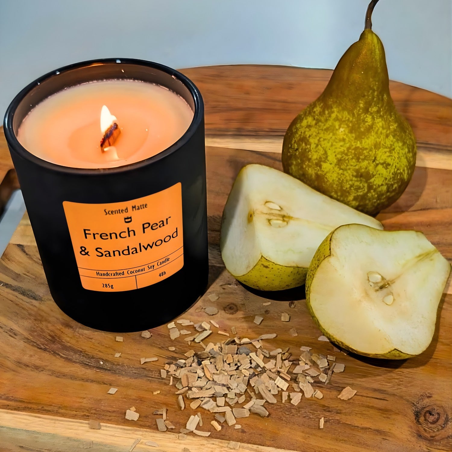 A minimalist matte black candle jar with a lit wooden wick, sitting next to a sliced green pear and light sandalwood chips.