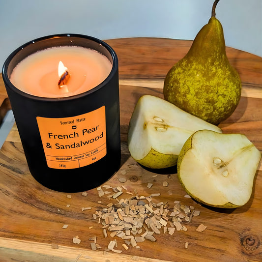 A minimalist matte black candle jar with a lit wooden wick, sitting next to a sliced green pear and light sandalwood chips.