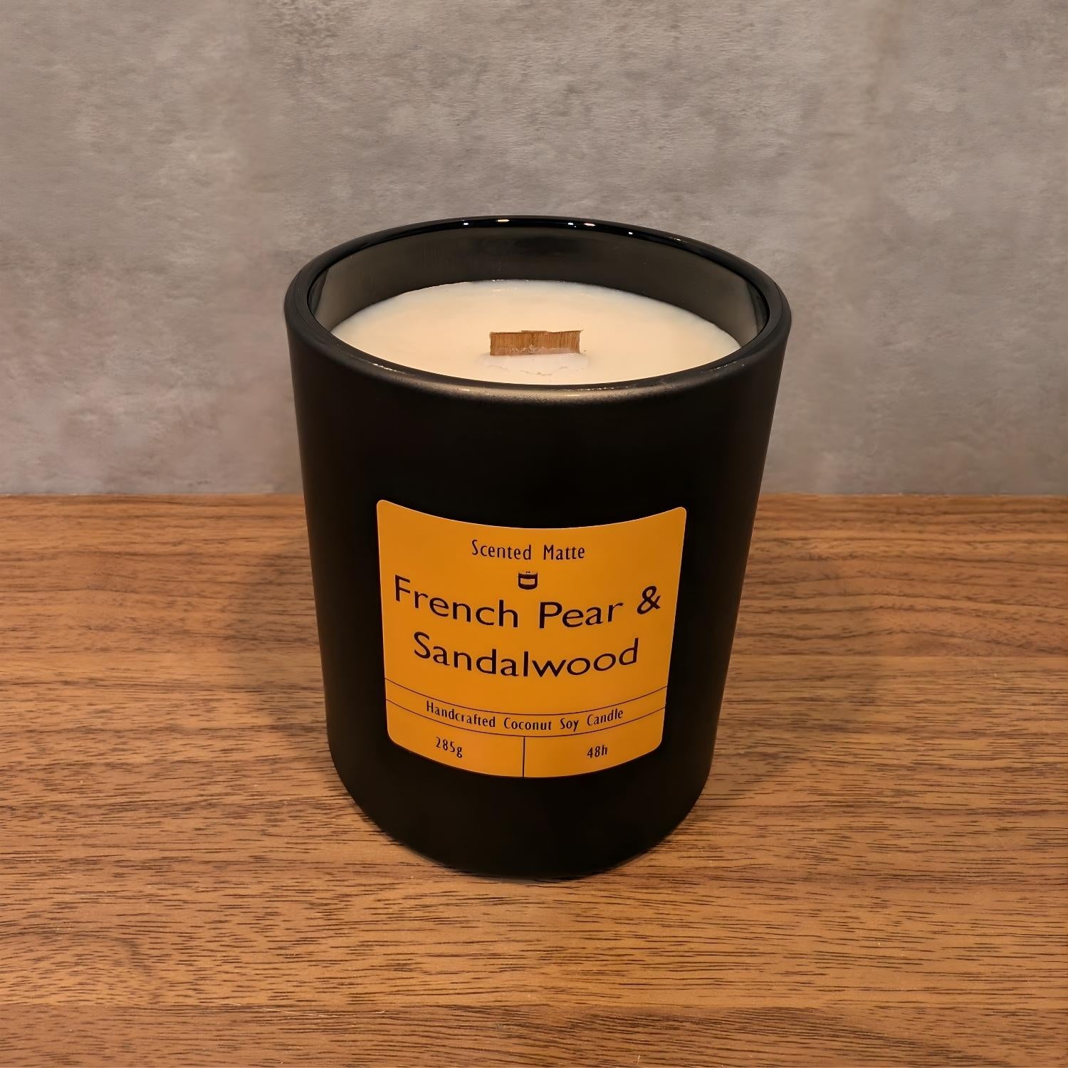 A minimalist matte black candle jar with a wooden wick, sitting on a light oak wood surface against a textured grey background.