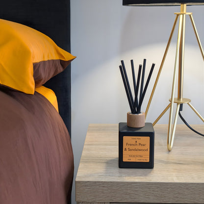 A minimalist matte black reed diffuser with orange label, wood cap and black reeds on a light wood bedside table, next to a gold tripod lamp and a bed with brown and mustard yellow linens.
