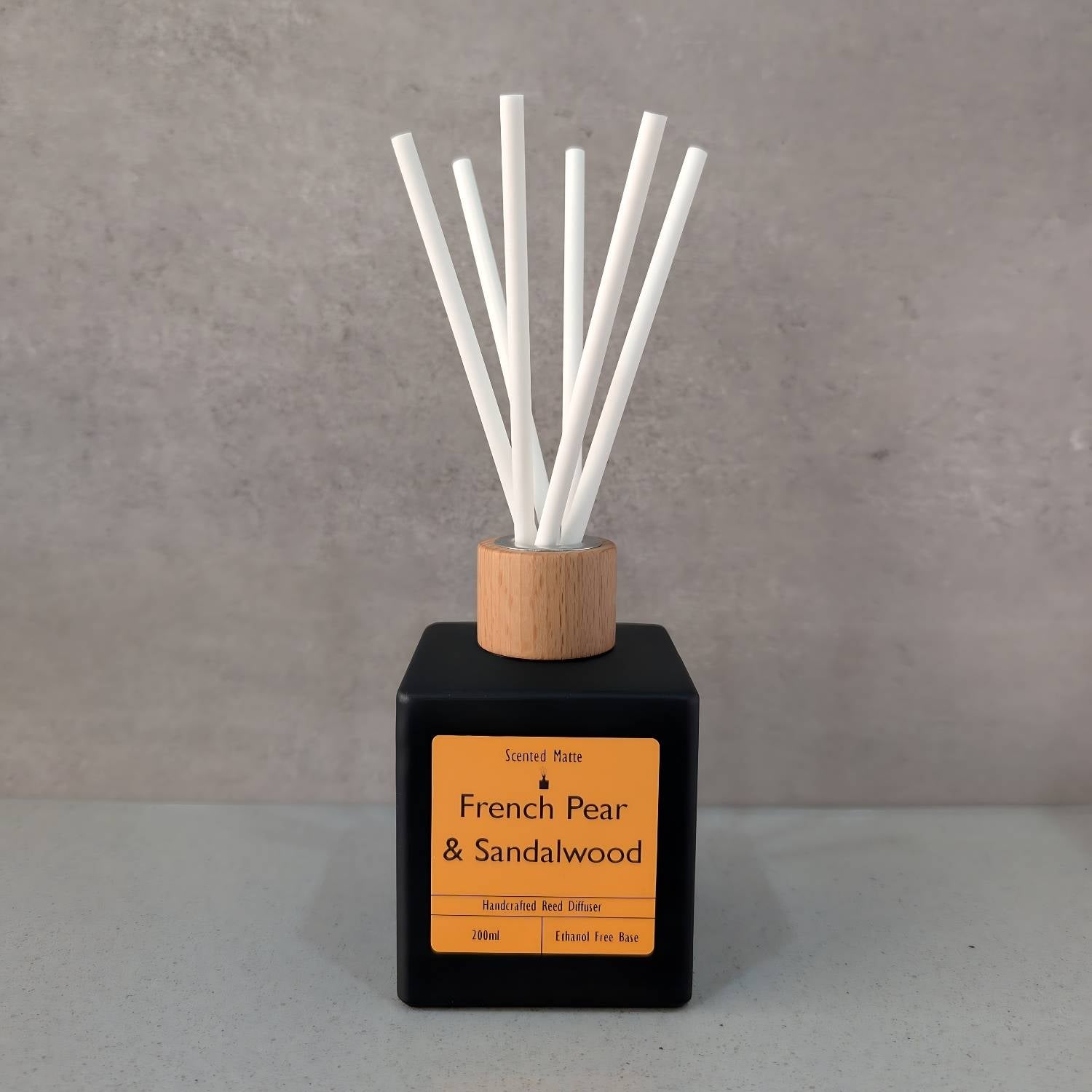 A minimalist matte black square reed diffuser with an orange label, light wood cap and six white fiber reeds, set against a grey background.