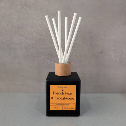 A minimalist matte black square reed diffuser with an orange label, light wood cap and six white fiber reeds, set against a grey background.