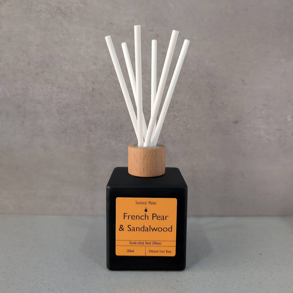 A minimalist matte black square reed diffuser with an orange label, light wood cap and six white fiber reeds, set against a grey background.