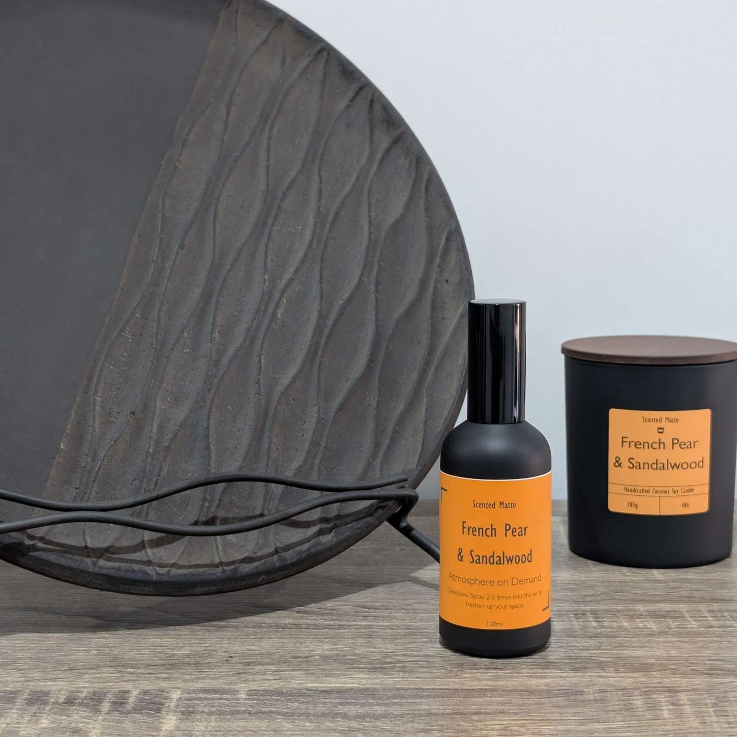 A matte black room spray bottle and a minimalist matte black candle jar, styled next to a large textured charcoal grey decorative plate.