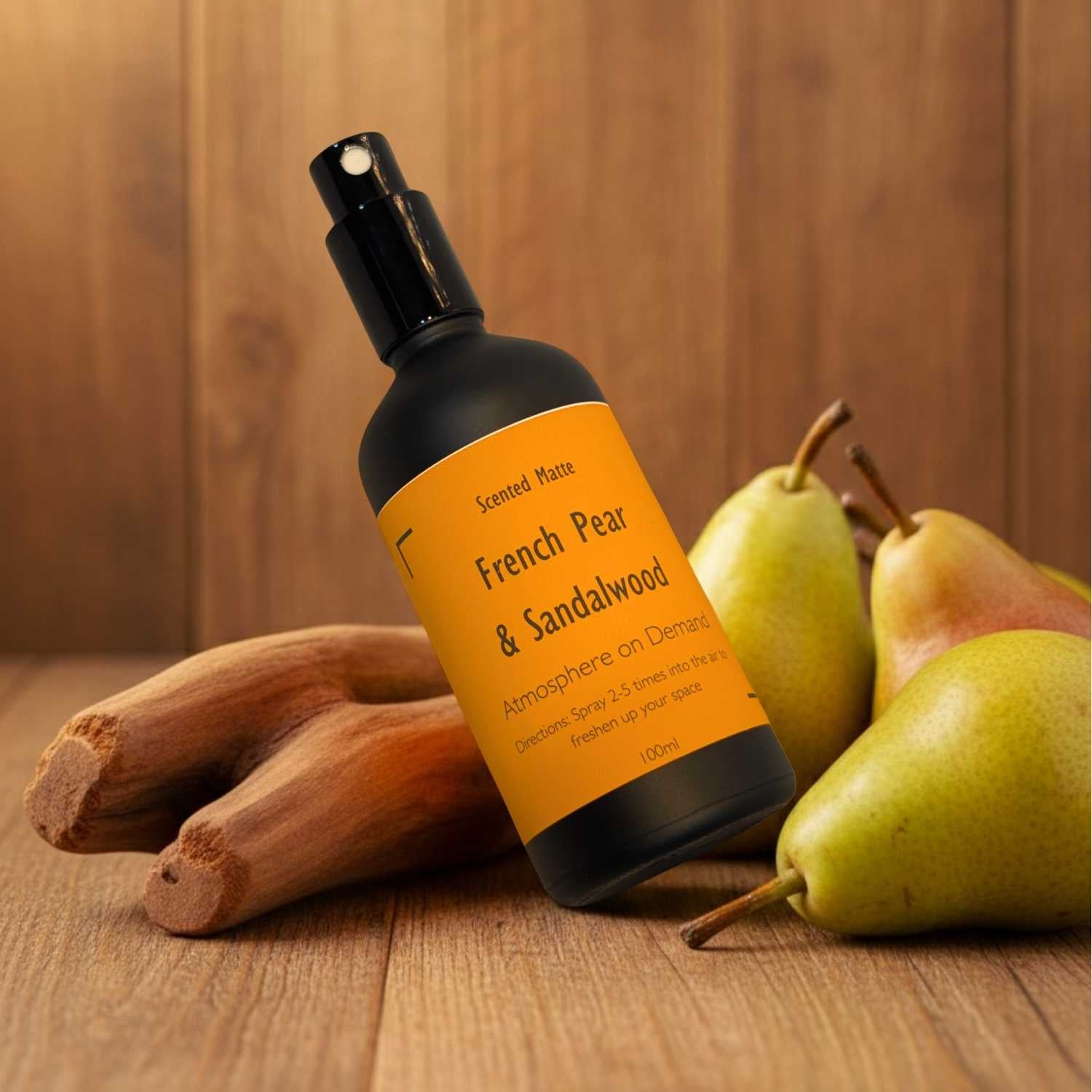 A minimalist matte black room spray bottle with a vibrant orange label, sitting next to a green pear and light sandalwood.
