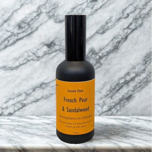 A minimalist matte black 100ml room spray bottle with a vibrant orange label, centered on a white marble surface.
