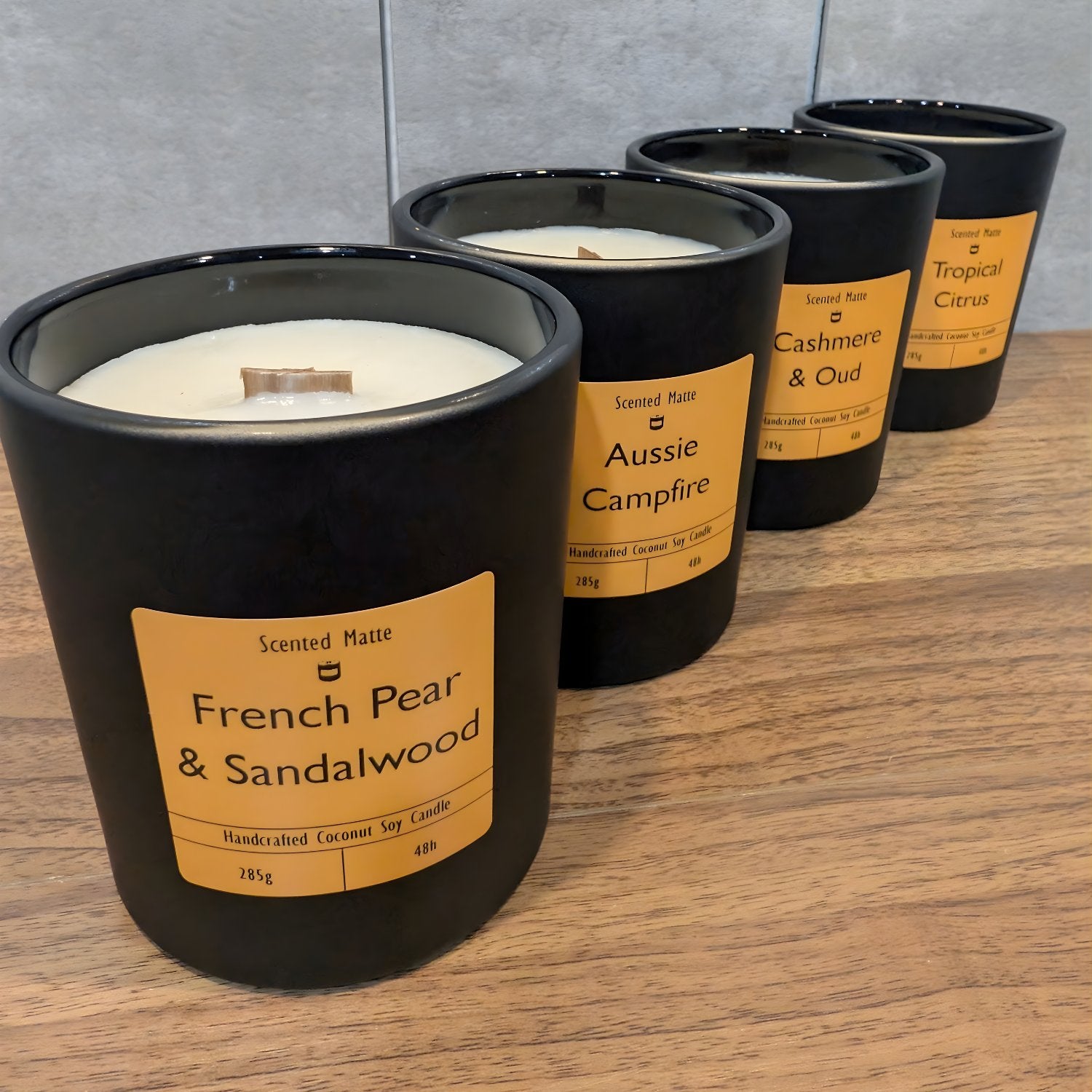 A high-angle diagonal row of four minimalist matte black jar candles with vibrant orange labels, staggered on a light wood surface against a grey tiled wall.