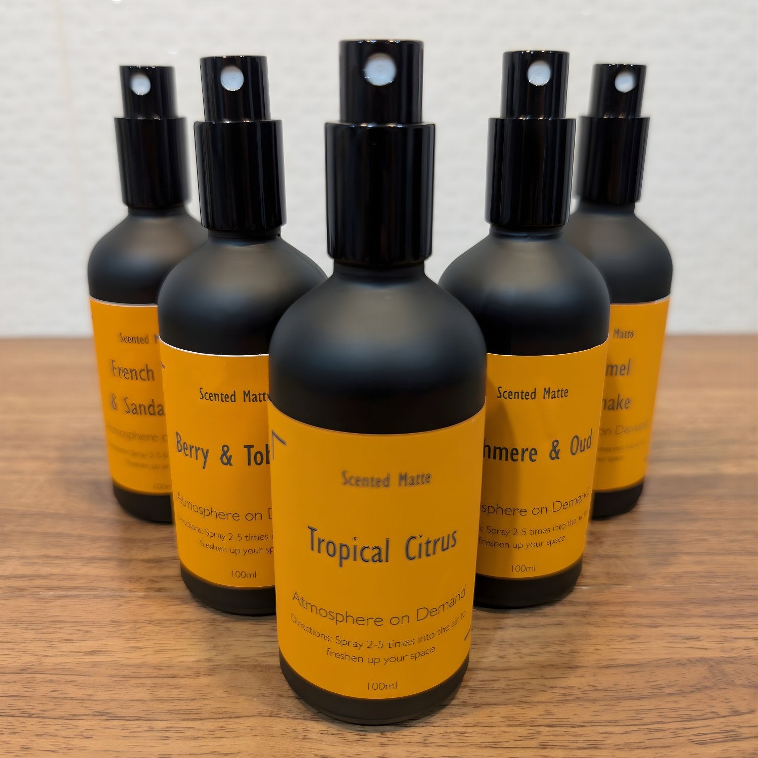 Set of five minimalistic matte black spray bottles with orange labels on a wooden surface.