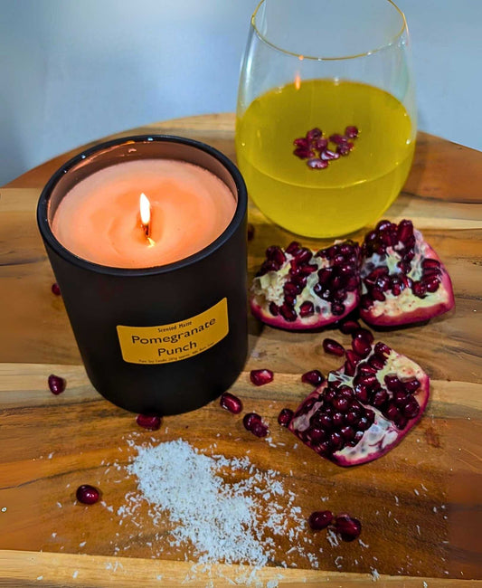 A round wooden board with a glass of yellow liquid with pomegranate seeds, pomegranate wedges and a lit wooden wick candle.