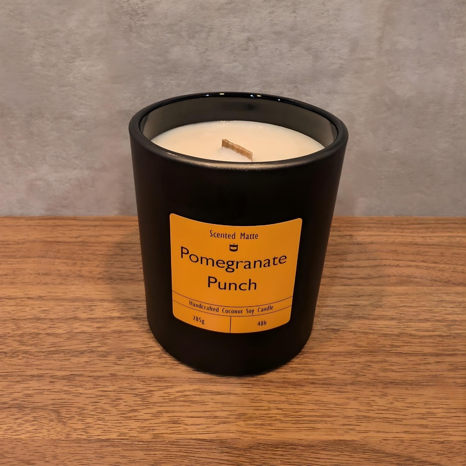 A minimalist matte black candle jar with a wooden wick, sitting on a light oak wood surface against a textured grey background.