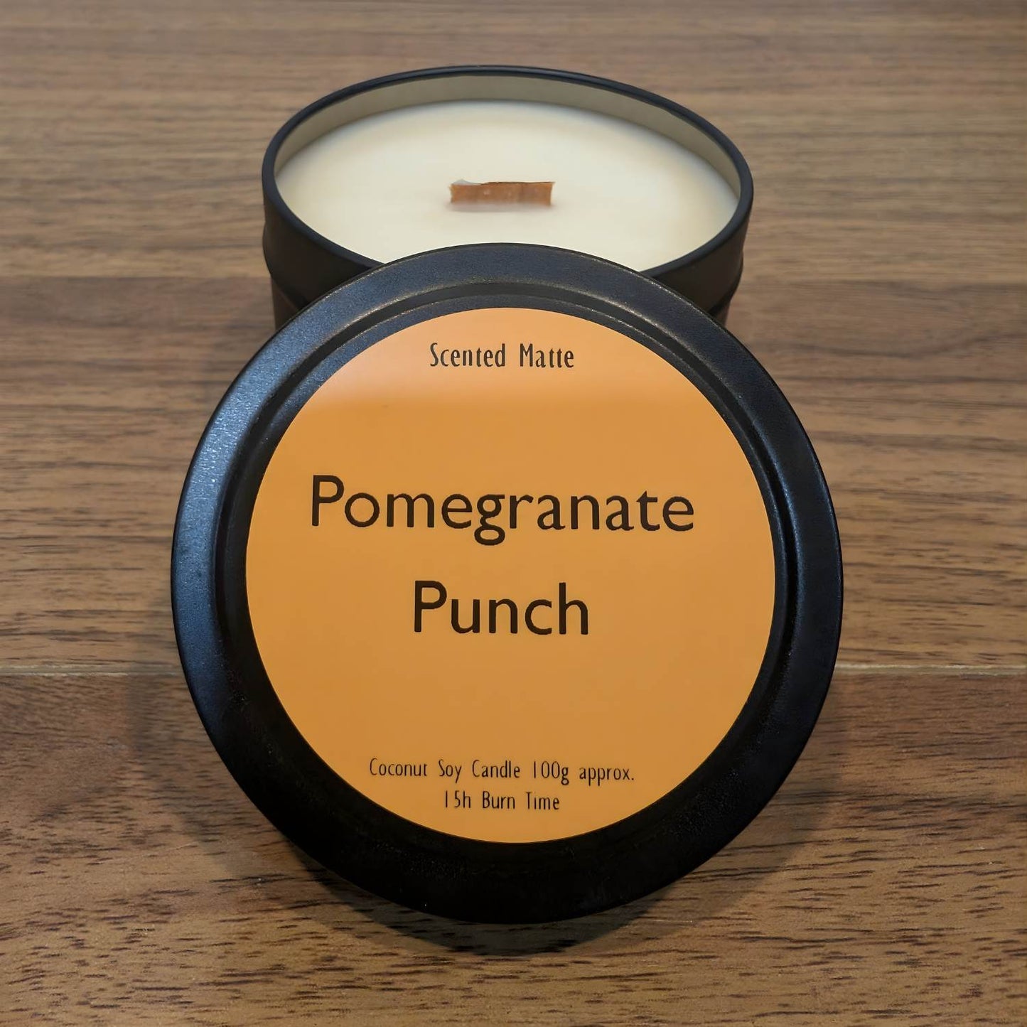 A wooden wick candle in a black tin with orange label on the lid on a wooden surface.
