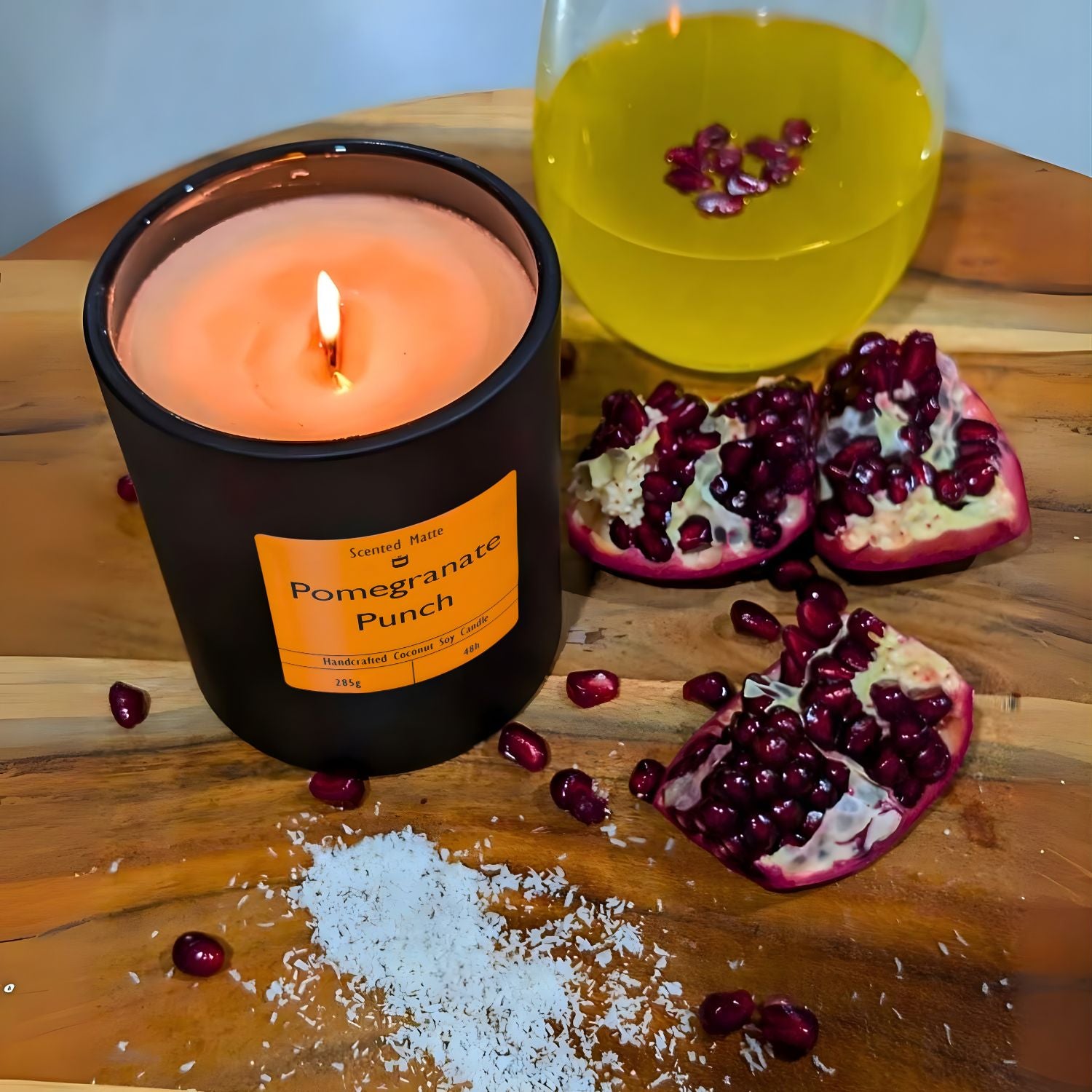 A minimalist matte black candle jar with a wooden wick, surrounded by ruby red pomegranate seeds and fresh citrus zest.